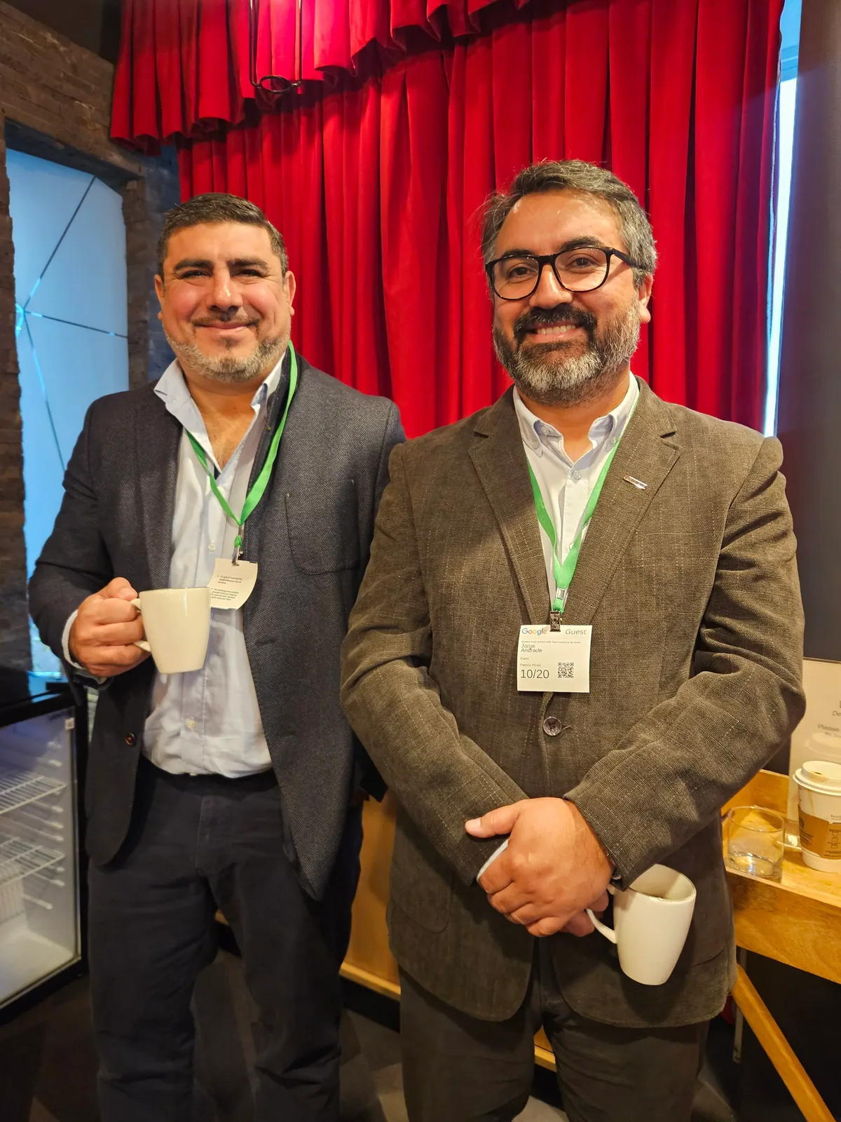 Dos asistentes sonríen con una taza de café frente a la cortina roja.